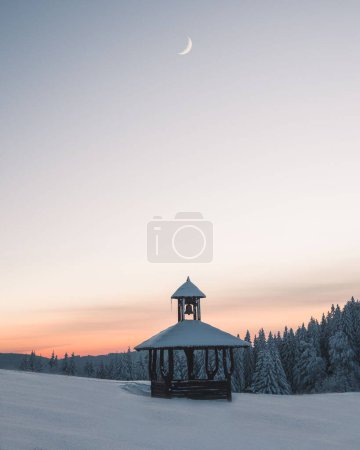 Foto de Un plano vertical de un edificio de madera con una campana cubierta de nieve bajo el cuarto de luna al atardecer - Imagen libre de derechos