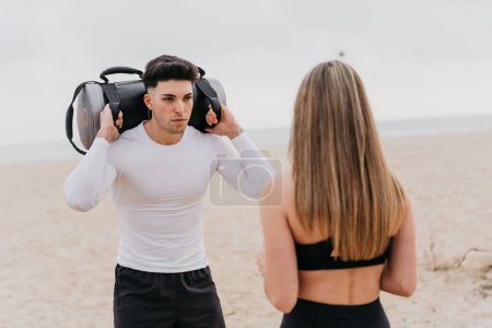 Téléchargez les photos : Un jeune homme athlétique avec une femme qui fait des squats à l'aide d'un sac de sable pour le poids - en image libre de droit