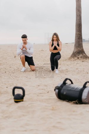 Photo for A young athletic couple exercising at the beach, doing squats with gym equipment - Royalty Free Image