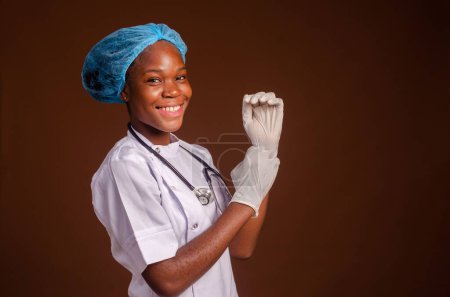 Téléchargez les photos : Une jeune infirmière noire heureuse ajustant des gants chirurgicaux isolés sur fond brun. - en image libre de droit