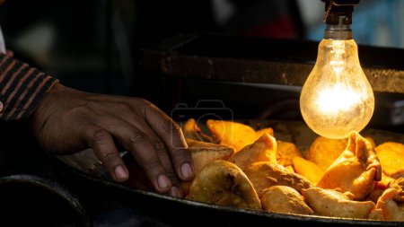 Téléchargez les photos : Gros plan de la main d'une personne ramassant un samosa sous une lampe d'éclairage. - en image libre de droit