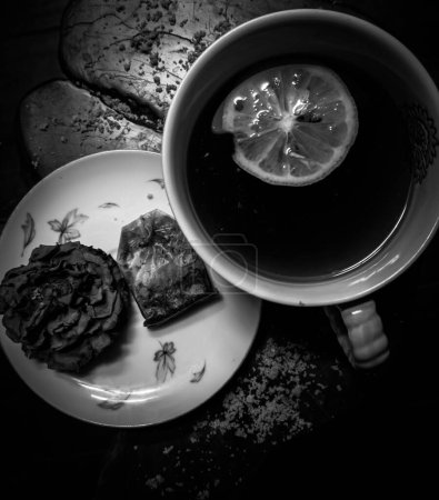 Photo for A top view grayscale of a cup of tea with lemon next to a plate with the tea bag - Royalty Free Image