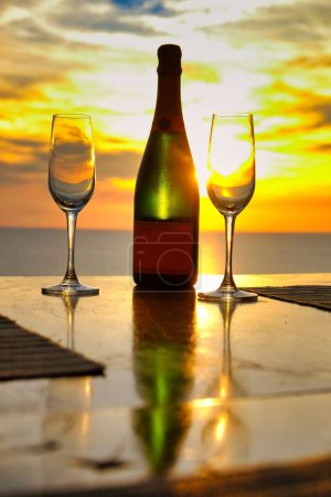 Foto de Una toma vertical de dos copas de vino y una botella en el fondo de una puesta de sol de color amarillo brillante - Imagen libre de derechos
