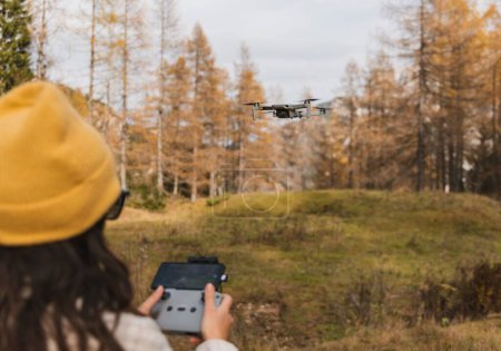 Foto de Vista trasera foto de enfoque selectivo de una mujer joven sosteniendo el mando a distancia mientras vuela un dron en el prado en las montañas - Imagen libre de derechos
