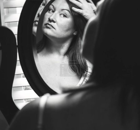 Téléchargez les photos : Une jeune femme caucasienne se regardant dans un miroir de vanité vérifiant ses cheveux et son maquillage, en niveaux de gris - en image libre de droit