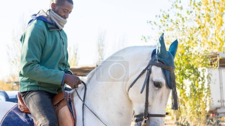 Foto de Un hombre negro adulto con ropa de abrigo montado en un caballo blanco en un rancho - Imagen libre de derechos