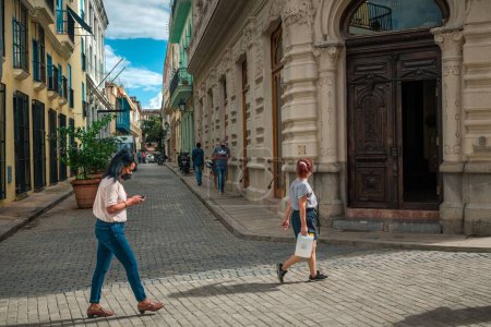 Foto de Los latinos cubanos caminando por La Habana restauraron edificios antiguos, el sol sobre los cuerpos de dos mujeres caminando y gente que venía de una calle estrecha. - Imagen libre de derechos