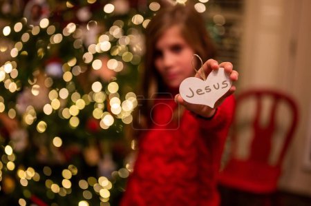 Photo for A girl holding a heart shaped paper with Jesus on it against the background of Christmas tree - Royalty Free Image