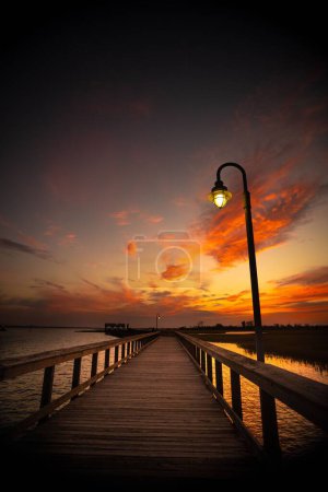 Photo for The wooden bridge over Shem Creek and a lamp under the orange-shaded sunset sky - Royalty Free Image