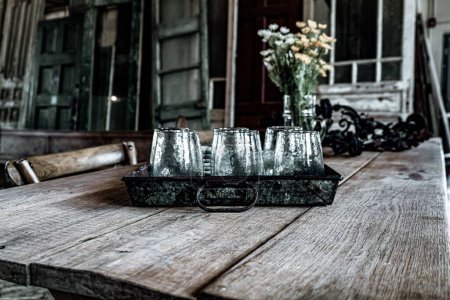 Photo for A Large wooden plank farmhouse table with glass cups in serving tray and distressed chairs - Royalty Free Image