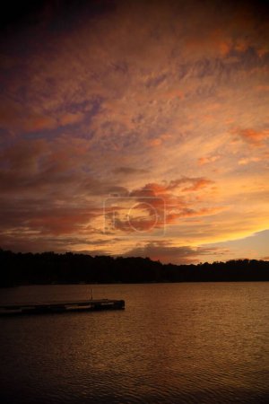 Téléchargez les photos : Une eau de lac calme entourée de hauts arbres verts contre un coucher de soleil pittoresque avec des nuages duveteux - en image libre de droit