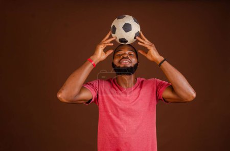 Téléchargez les photos : Un homme africain heureux attrayant tenant un ballon de football isolé sur un fond brun. - en image libre de droit