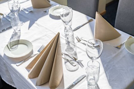 Téléchargez les photos : Une table à manger vide avec des tasses en verre, cuillères, fourchettes, couteaux et serviettes ensoleillées dessus - en image libre de droit