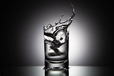 Foto de Un primer plano a escala de grises de vaso de agua con salpicaduras de cubo de hielo cayendo. - Imagen libre de derechos