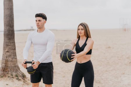 Foto de Una joven pareja atlética haciendo ejercicio en la playa con un kettlebell y una pelota de medicina - Imagen libre de derechos