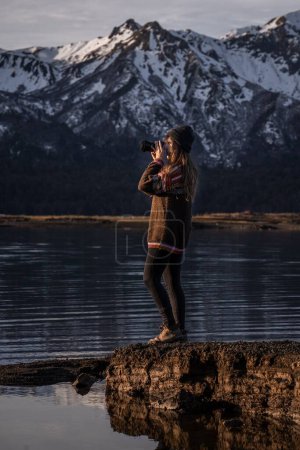 Téléchargez les photos : Prise de vue verticale d'une femme debout sur le rivage et photographiant la montagne enneigée dans les hautes terres - en image libre de droit