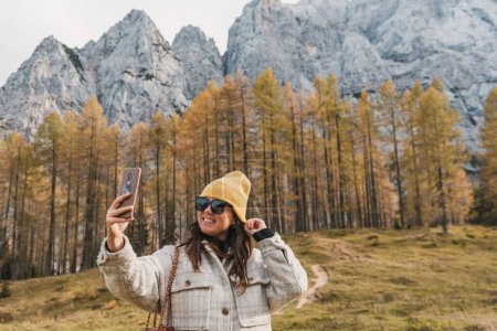 Photo for Portrait of happy young woman taking a selfie in front of golden larch trees under mountains in autumn at Vrsic mountain pass in Slovenia. - Royalty Free Image