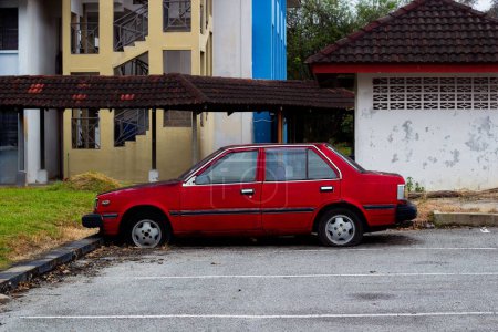 Téléchargez les photos : Une vieille voiture Proton Saga abandonnée rouge garée dans la cour près des maisons - en image libre de droit