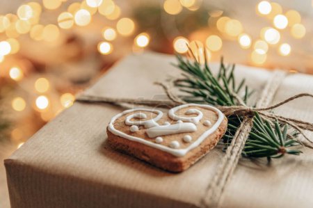 Téléchargez les photos : Photo en gros plan de biscuit au pain d'épice en forme de cœur à Noël présent devant les lumières bokeh. Cadeau de Noël enveloppé dans du papier brun et décorer - en image libre de droit