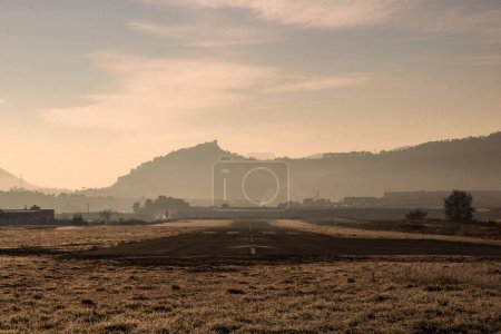 Téléchargez les photos : Une piste d'aéroport avec des panneaux de direction entourés de terrains herbeux et des collines avec des arbres au coucher du soleil - en image libre de droit