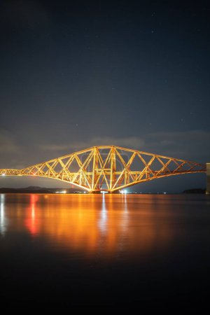 Téléchargez les photos : Beaux paysages du célèbre pont ferroviaire de Forth éclairé par des veilleuses réfléchies dans l'eau en Écosse, au Royaume-Uni, sous un ciel étoilé - en image libre de droit