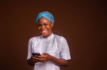 Téléchargez les photos : Un beau portrait d'un jeune médecin heureux textant avec un smartphone sur un fond brun - en image libre de droit