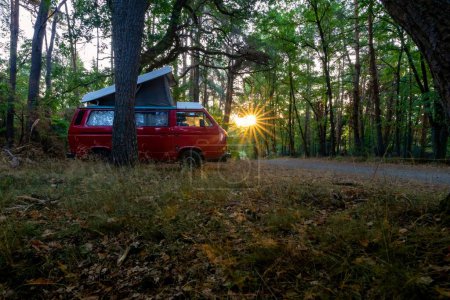 Téléchargez les photos : Un camping-car rouge garé sur la route principale dans une forêt avec des rayons de soleil couchant à travers les arbres verts en arrière-plan - en image libre de droit