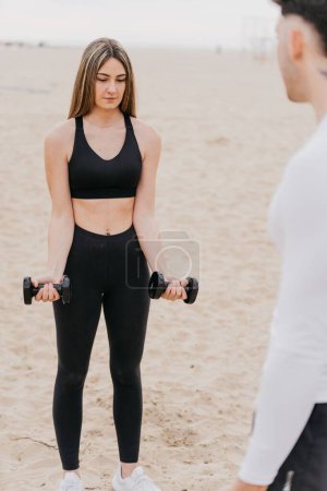 Foto de Una foto vertical de una atlética en forma haciendo rizos de bíceps usando pesas con un entrenador masculino en la playa - Imagen libre de derechos