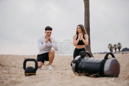 Foto de Una joven pareja atlética haciendo ejercicio en la playa, haciendo sentadillas con equipo de gimnasio - Imagen libre de derechos