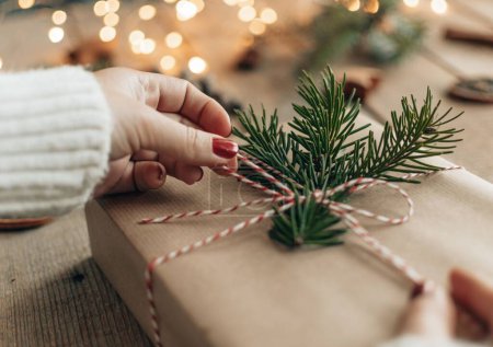 Téléchargez les photos : Photo gros plan de la main féminine avec vernis à ongles rouge, enveloppant ou ouvrant cadeau de Noël enveloppé dans du papier brun et décoré avec une brindille d'épinette verte. - en image libre de droit