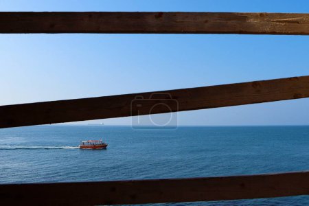 Photo for A view through a fence of a red boat sailing on the blue water of Acre harbor, Israel - Royalty Free Image