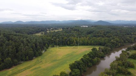 Téléchargez les photos : Une vue aérienne de la campagne avec un champ vert entouré de forêts denses luxuriantes à côté d'une rivière étroite - en image libre de droit