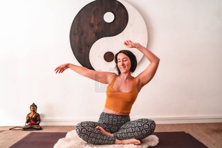 Foto de Una joven hembra en forma haciendo yoga en una casa alfombra cerca de una decoración de pared yin yang - Imagen libre de derechos