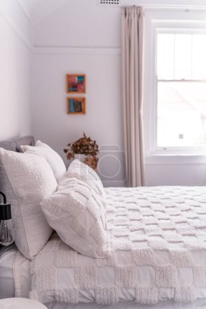 Foto de El enfoque selectivo de las cuatro almohadas blancas y la cubierta de la cama con dibujos al lado de las imágenes de la pared, una planta seca y una ventana - Imagen libre de derechos