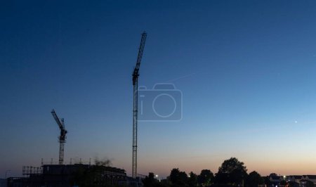 Téléchargez les photos : Vue à angle bas des grues à silhouette au-dessus du bâtiment et des arbres contre un ciel bleu clair au coucher du soleil - en image libre de droit
