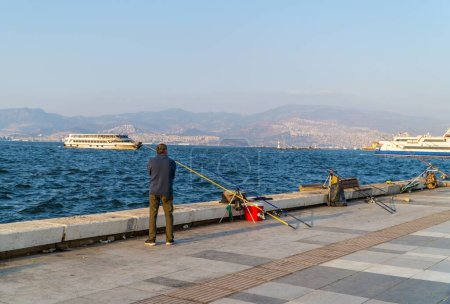 Téléchargez les photos : Un pêcheur pêche par une journée ensoleillée sur la côte de la baie d'Izmir, Turquie, avec des navires et une montagne en arrière-plan - en image libre de droit