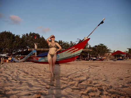 Téléchargez les photos : Une jolie femme caucasienne en maillot de bain posant sur la plage de sable avec un bateau en arrière-plan - en image libre de droit