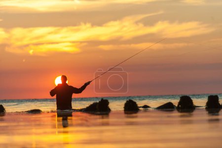 Téléchargez les photos : Vue arrière silhouette de l'homme pêchant avec la canne tout en se tenant en mer contre le ciel nuageux pendant le coucher du soleil - en image libre de droit