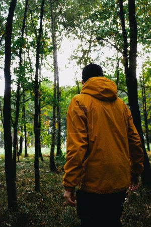Photo for A vertical back view of the traveler with an orange coat walking in the lush forest - Royalty Free Image