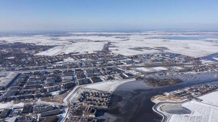 Téléchargez les photos : Vue aérienne d'une ville, avec une rivière gelée et des champs enneigés par une journée d'hiver nuageuse - en image libre de droit