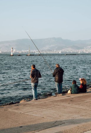 Téléchargez les photos : Vue verticale des pêcheurs pêchant par une journée ensoleillée sur la côte de la baie d'Izmir, en Turquie, et une montagne brumeuse en arrière-plan - en image libre de droit
