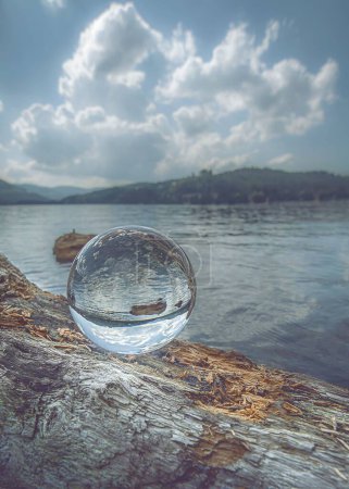 Foto de Un disparo vertical de una bola de cristal sobre un tronco de árbol caído, que refleja un lago tranquilo en un día nublado - Imagen libre de derechos