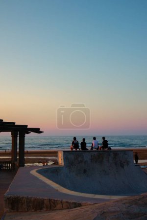 Foto de Un tiro de espalda de hombres jóvenes sentados con sus patinetas en el borde de un patio de skate en la playa al atardecer - Imagen libre de derechos