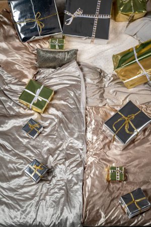 Photo for High angle view of gift boxes wrapped with silver and golden papers arranged on messy sheets on bed at home - Royalty Free Image