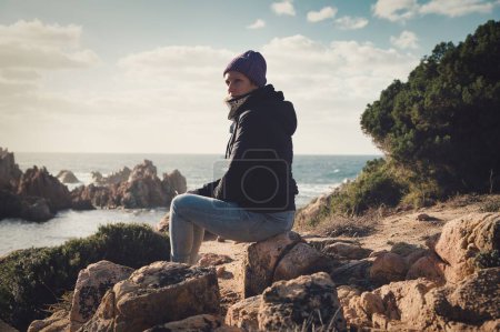 Photo for Attractive woman dressed in winter clothes sitting by the sea - Royalty Free Image