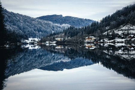 Téléchargez les photos : Une maison au bord du lac contre une montagne couverte d'arbres en hiver avec réflexion dans l'eau - en image libre de droit
