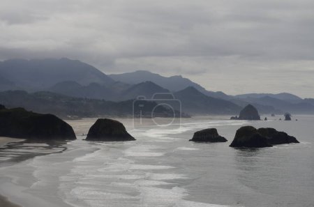 Téléchargez les photos : Une vue sur le paysage des rochers de la plage Canon en Oregon avec des collines par une journée nuageuse - en image libre de droit