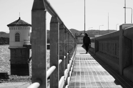 Téléchargez les photos : Plan en niveaux de gris d'une personne marchant sur un pont près d'un phare à la lumière du jour - en image libre de droit