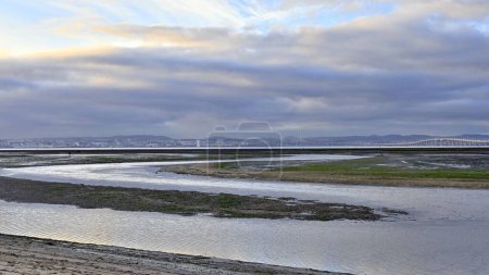 Téléchargez les photos : Le bord de mer à marée basse sur fond de bâtiments de la ville et pont sur ciel nuageux - en image libre de droit