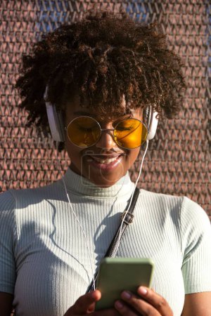 Foto de Una joven latina con cabello afro y gafas sonriendo usando un smartphone para redes sociales - Imagen libre de derechos